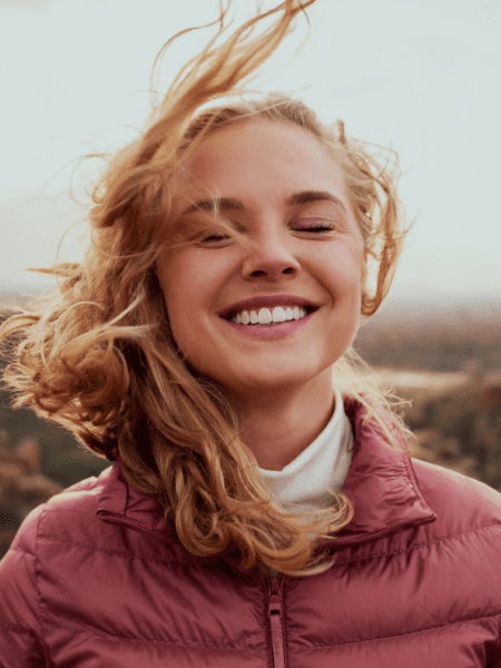 Imagem de mulher jovem, cabelos loiros, sorrindo de olhos fechados, ao ar livre, demonstrando paz. Imagem de fundo na pagina cadore spa